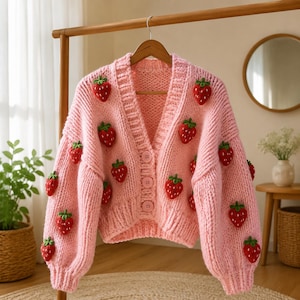Può includere: Un cardigan in maglia grossa di colore rosa pastello tenue con scollo a V, maniche lunghe e chiusura frontale con bottoni. Il capo è decorato con molteplici applicazioni tridimensionali a forma di fragola rossa con foglie verdi.