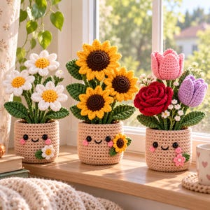 May include: Three crocheted potted plants featuring white daisies, yellow sunflowers, and a mix of red, pink, and purple flowers. Each beige pot has a stitched smiling face with black eyes and pink cheeks, decorated with a small crocheted flower.