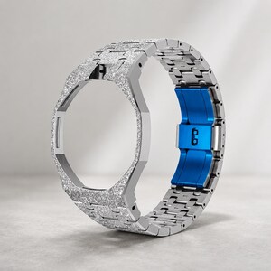 Könnte beinhalten: Ein silberfarbenes Uhrengehäuse mit integriertem Gliederarmband, das eine texturierte, glitzernde Oberfläche auf den Außenseiten aufweist. Der innere Verschlussmechanismus ist in einem leuchtenden, einfarbigen Blau gehalten.
