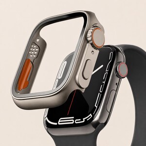 Könnte beinhalten: Ein metallisch graues Schutzgehäuse für eine Uhr, das über einer dunklen Smartwatch mit schwarzem Silikonarmband schwebt. Das Gehäuse hat eine seitliche Krone mit orangefarbenem Ring, während die Uhr ein digitales Zifferblatt zeigt.