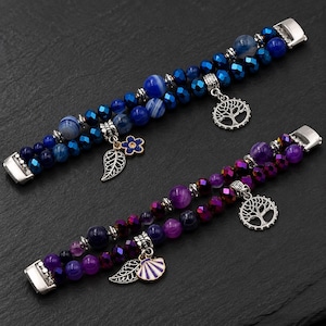 Op de afbeelding: Twee meerstrengs armbanden met gepolijste kralen in blauwe en paarse tinten. De blauwe armband bevat een zilveren boombedel en een blauwe bloem met een bladbedel. De paarse armband heeft een zilveren boombedel en een schelpbedel.