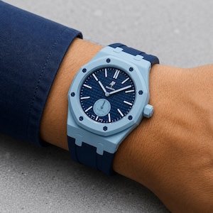 Könnte beinhalten: Eine hell- und dunkelblaue Armbanduhr mit achteckiger Lünette und acht sichtbaren Schrauben. Das dunkelblaue Zifferblatt hat ein strukturiertes Gittermuster, weiße Stundenmarkierungen und den Schriftzug AP Swatch Royal Oak. Mit dunkelblauem Silikonarmband.