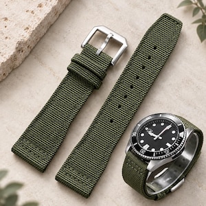 Peut inclure: Un bracelet de montre en tissu tissé vert olive avec une boucle en métal argenté et une montre assortie dotée d'un cadran noir, d'index blancs et d'une lunette rotative avec des marquages argentés, présenté en deux parties distinctes.