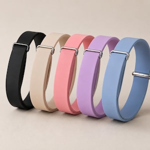 Può includere: Cinque cinturini in silicone regolabili nei colori nero, beige, rosa, lavanda e azzurro. Ogni cinturino presenta una finitura opaca liscia e una fibbia scorrevole in metallo argentato per regolare la lunghezza, disposti in fila su una superficie neutra.
