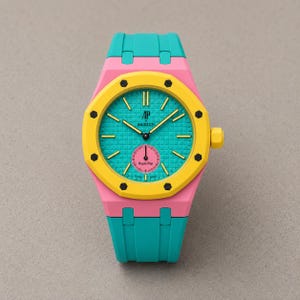 Puede incluir: Un reloj de pulsera con un bisel octogonal amarillo brillante con ocho tornillos negros, una esfera texturizada turquesa con los logotipos AP y Swatch, y una subesfera rosa con la etiqueta Royal Pop. El reloj tiene una caja rosa y correa turquesa.