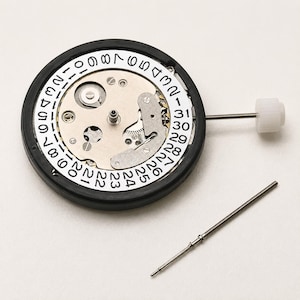 May include: A circular watch movement mechanism featuring a white date wheel with black numerals from 1 to 31. The metallic internal components are visible, surrounded by a black ring. A white crown is attached to a stem, with a separate metal stem nearby.