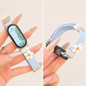 Puede incluir: Una correa de reloj trenzada de color azul claro con un detalle de flor de margarita blanca y amarilla. La correa está unida a un rastreador de actividad digital ovalado con una pantalla floral que muestra la hora 09:28 y la fecha en tonos azul y verde.