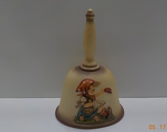 May include: A ceramic bell with a white handle and a painted image of a girl in a red and white dress holding a leaf. The bell is decorated with brown, red, white, and green accents.