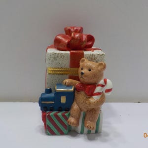 May include: A Christmas-themed figurine featuring a brown teddy bear holding a candy cane and wearing a red bow. The bear sits atop a stack of presents and a blue train. The presents are white with red stripes and a red bow.