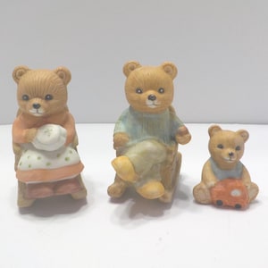 May include: Three ceramic teddy bear figurines. One bear is seated in a chair and wearing an orange dress and a white bonnet. Another bear is seated in a chair and wearing a blue sweater. The third bear is holding a small orange car.