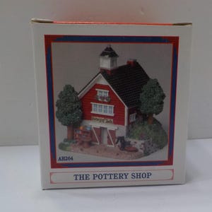 May include: A decorative miniature of "The Pottery Shop" in a box. The shop is a red barn with a black roof, white trim, and two green trees. The box has a red and blue border and the text "THE POTTERY SHOP".