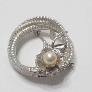 May include: A silver tone brooch with a faux pearl in the center of a flower design. The flower is surrounded by a circle of twisted silver.