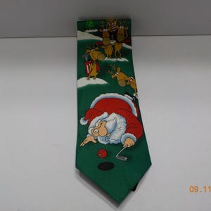 May include: A green necktie featuring a cartoon illustration of Santa Claus in a red suit playing golf on a snowy course. Above Santa, several brown reindeer are depicted in various poses, including some wearing scarves and holding golf clubs.