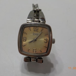 May include: A silver-toned wristwatch with a square face and a light brown dial. The watch has a silver band and a small second hand. The face of the watch has black Roman numeral hour markers and the word "Ice" printed on the dial.