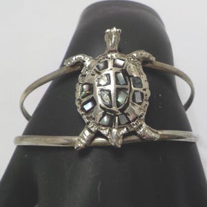 May include: A silver-toned bracelet featuring a turtle design. The turtle's shell is inlaid with iridescent pieces, and the bracelet has a simple, open design. The bracelet is displayed on a black form.