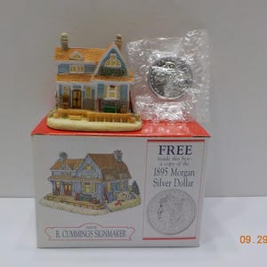 May include: A miniature house figurine with a blue and yellow roof, sitting on a red box. The box features an illustration of the house and text that reads "FREE inside this box - a copy of the 1895 Morgan Silver Dollar."