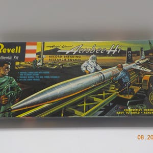 May include: A Revell Aerobee-Hi research rocket model kit box featuring a silver rocket on a launch trailer with crew figures. The box displays the text Aerobee-Hi, Record-Breaking Research Rocket, and lists features like movable firing arm and wheels.