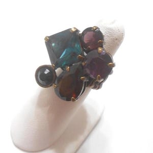 May include: A cluster ring featuring a variety of colorful gemstones. The ring has a bronze-colored band and is adorned with a large, square teal stone, several round purple stones, and a smaller black stone. The gemstones are set in a cluster design.