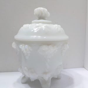 May include: A white, lidded jar with a decorative grape design. The jar has a rounded body, three small feet, and a knob on the lid. The jar is made of opaque white glass.