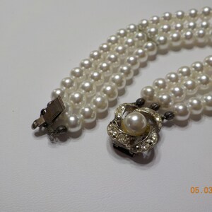 May include: A three-strand faux pearl necklace with a silver clasp and a decorative silver pendant featuring a faux pearl surrounded by rhinestones.