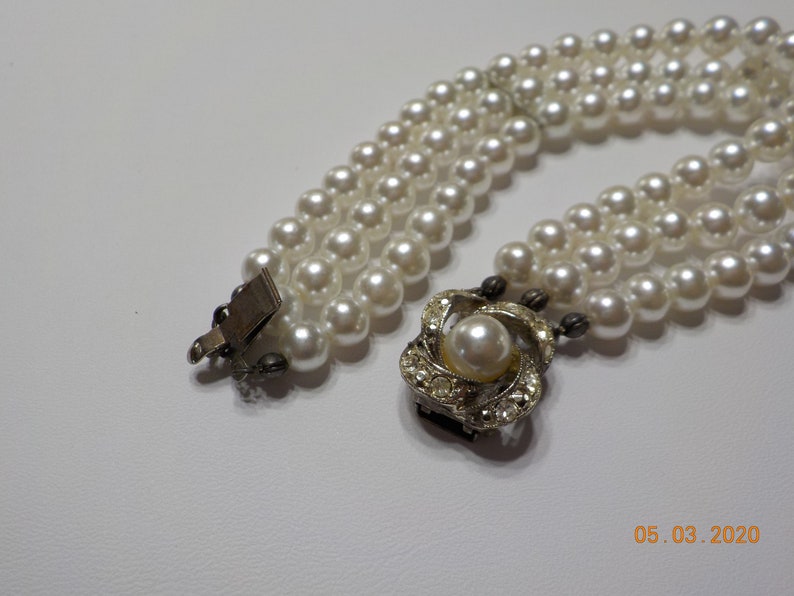 May include: A three-strand faux pearl necklace with a silver clasp and a decorative silver pendant featuring a faux pearl surrounded by rhinestones.
