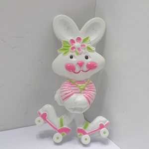 May include: A white bunny figurine with pink cheeks, a pink and green flower crown, and a pink and white striped shirt. The bunny is wearing roller skates with pink and green accents.