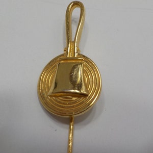May include: A gold-toned metal pin with a circular design featuring a bell-shaped charm. The pin has a long, thin stem.