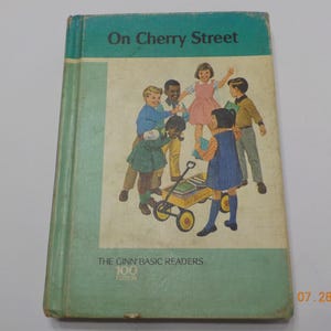 May include: Vintage children's book titled "On Cherry Street." The cover features illustrations of children around a wagon. The book is part of "The Ginn Basic Readers" series, 100th edition. The cover is teal and cream colored.