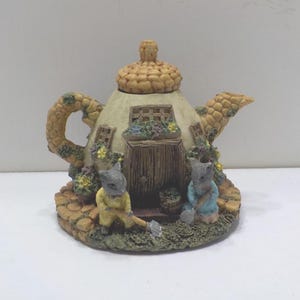 May include: A whimsical teapot-shaped figurine with a cottage design. The beige teapot features a textured lid and spout, a wooden door, and two small figures. The base is a stone-like texture, and the scene is adorned with flowers.