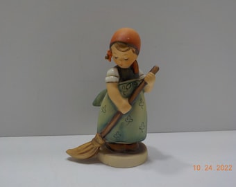 May include: A figurine of a girl in a green dress and red hat holding a broom. She is looking down at the broom and has a sweet expression on her face.