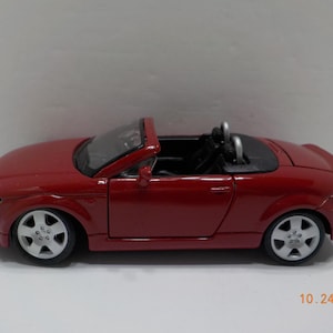 May include: A red toy car model of an Audi TT Roadster convertible. The car has black interior and silver wheels.