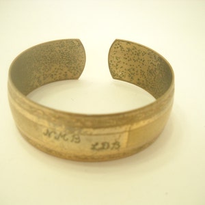 May include: A vintage brass cuff bracelet with an open design. The bracelet has a textured surface and a band with engraved text. The overall color is a warm, aged gold.
