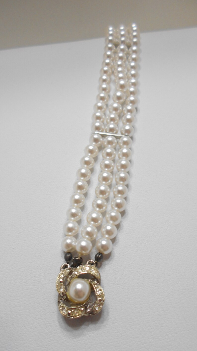 May include: A triple strand pearl bracelet with a decorative clasp featuring a pearl surrounded by rhinestones.