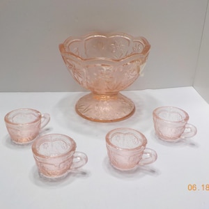 May include: A vintage pink glass dessert set featuring a large bowl with a pedestal base and four small handled cups. The set has a floral design and is ideal for serving desserts or small portions.