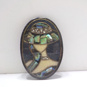 May include: Oval-shaped vintage brooch featuring a woman's silhouette. The brooch has a dark blue background with iridescent abalone shell accents. The woman's hat and neckline are adorned with rhinestones, and the face is a cream color.