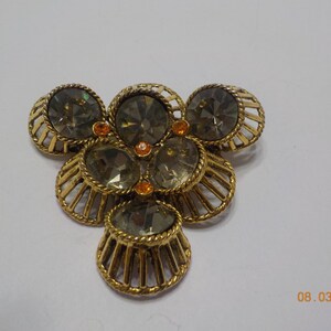 May include: A vintage brooch with a floral design. The brooch features gold-toned metalwork and is adorned with smoky gray and orange-colored rhinestones. The design is symmetrical and detailed.