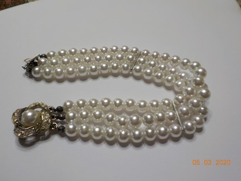 May include: A triple strand pearl necklace with a silver clasp and a decorative silver and rhinestone pendant.