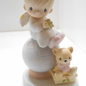 May include: A porcelain figurine of an angel with blonde hair and a halo, sitting on a globe with a teddy bear at her feet. The angel is wearing a white dress and holding a green leaf. The teddy bear is wearing a pink bow.
