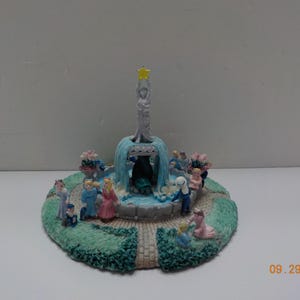 May include: A miniature fountain sculpture with a central figure holding a star. The fountain has a blue waterfall effect, surrounded by small figures and floral accents. The base is a circular garden design.