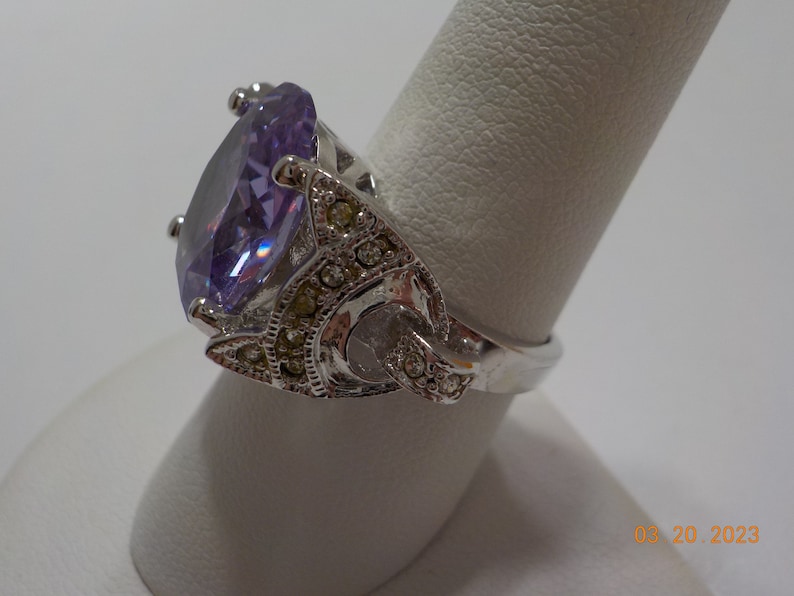 May include: A silver ring with a large, oval-shaped, purple gemstone surrounded by smaller, clear gemstones.