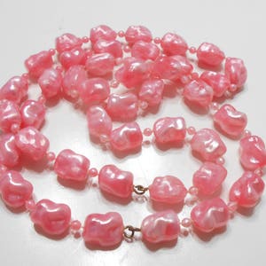 May include: A long pink pearl necklace with irregular shaped beads. The necklace is made of plastic and has a shiny finish.