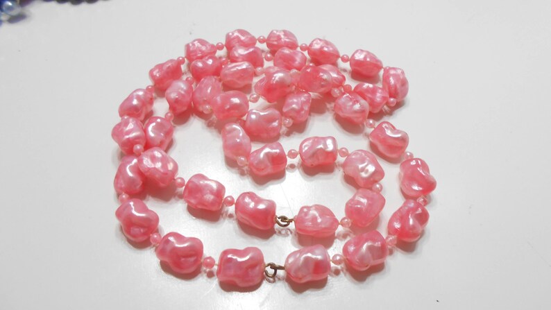 May include: A long pink pearl necklace with irregular shaped beads. The necklace is made of plastic and has a shiny finish.