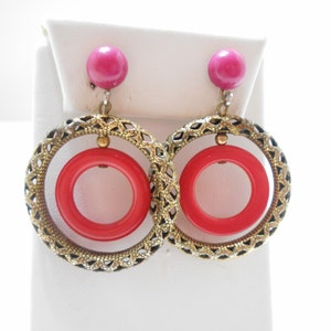May include: A pair of dangle earrings with a gold-toned metal frame and a bright red circle pendant. The top of the earring features a small pink bead.