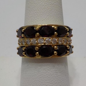 May include: A gold ring with three rows of gemstones. The top and bottom rows are red oval-shaped gemstones, and the middle row is clear round gemstones.
