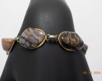 May include: A bracelet featuring polished, irregularly shaped stones in shades of brown, gray, and cream. The stones are set in gold-toned metal with a textured, scalloped edge, connected by silver-toned links. A unique piece of jewelry.