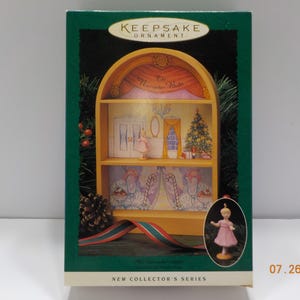 May include: A Keepsake Ornament box featuring a yellow, arched, two-shelf display unit with a Nutcracker Suite theme. The interior displays a Christmas tree, doors, and candy cane illustrations. A small figurine in a pink dress is shown in an oval inset.