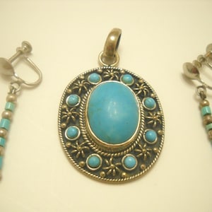 Op de afbeelding: Een turquoise hangerketting en bijpassende oorbellen met een zilverkleurige metalen setting en een turquoise steen. De hanger heeft een cirkelvormig ontwerp met een grote ovale turquoise steen in het midden.