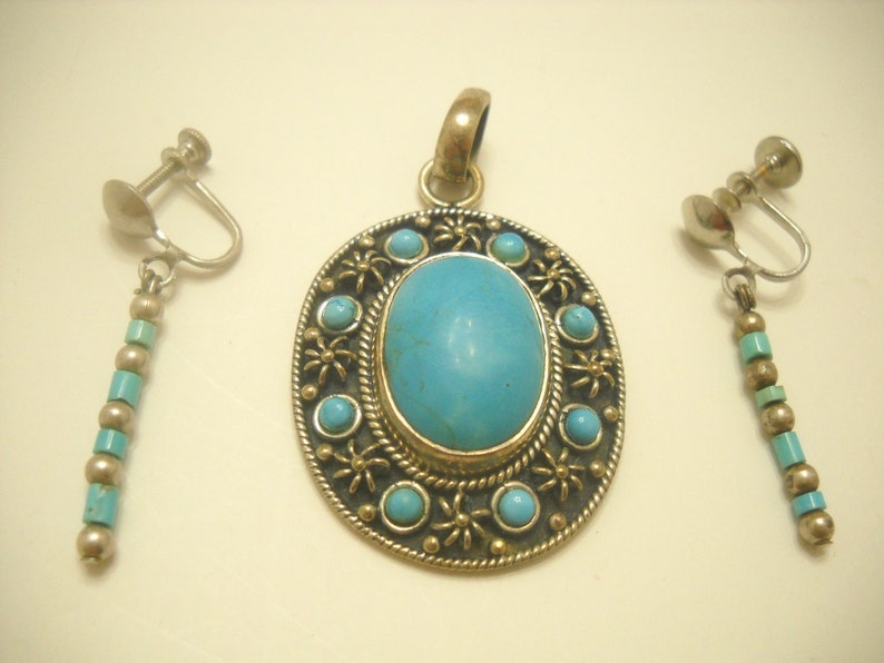 Op de afbeelding: Een turquoise hangerketting en bijpassende oorbellen met een zilverkleurige metalen setting en een turquoise steen. De hanger heeft een cirkelvormig ontwerp met een grote ovale turquoise steen in het midden.