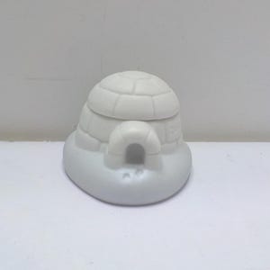 May include: A small, white igloo-shaped decorative item. The igloo has a rounded top with a segmented design and a curved entrance. The base is oval-shaped, and the overall appearance is smooth and clean.