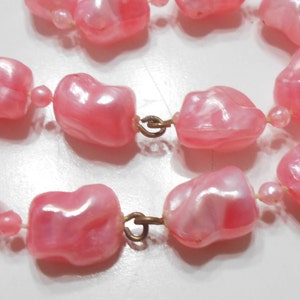 May include: A pink plastic pearl necklace with a gold clasp. The pearls are irregular in shape and have a slight iridescent sheen.